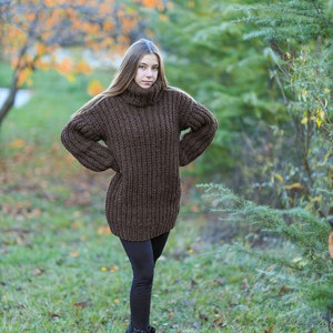 May include: A woman wearing a brown, oversized turtleneck sweater. The sweater is a chunky knit and reaches to the mid-thigh. She is also wearing black leggings and black sneakers with white soles. The background is a park with trees and grass.