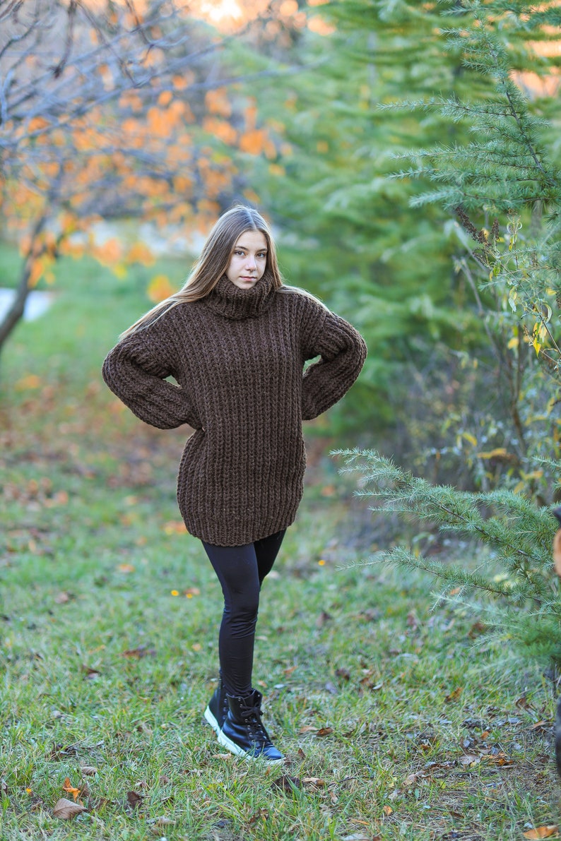 May include: A woman wearing a brown, oversized turtleneck sweater. The sweater is a chunky knit and reaches to the mid-thigh. She is also wearing black leggings and black sneakers with white soles. The background is a park with trees and grass.