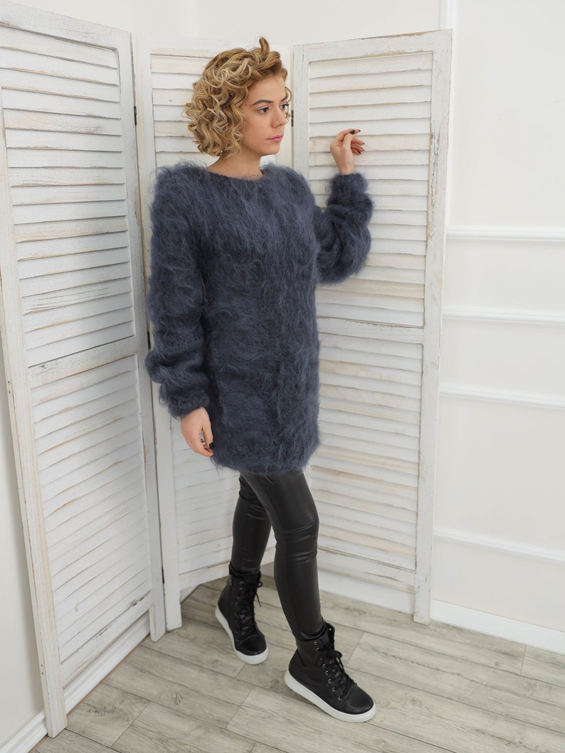 May include: A long, fuzzy, dark blue sweater with long sleeves. The sweater is worn by a person with curly blonde hair and black leggings. The person is standing in front of a white wooden screen.
