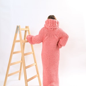 May include: A person wearing a long, pink knitted sweater dress. The sweater is very chunky and has a high neck. The person is standing next to a wooden step ladder.