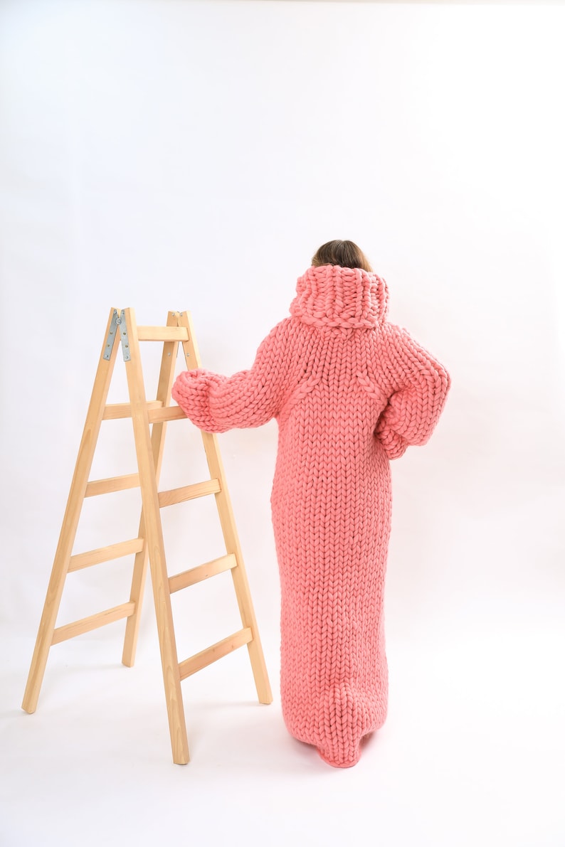May include: A person wearing a long, pink knitted sweater dress. The sweater is very chunky and has a high neck. The person is standing next to a wooden step ladder.