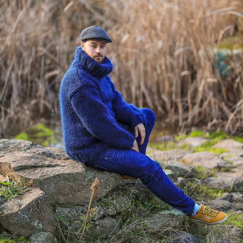 May include: A man wearing a blue turtleneck sweater and blue jeans sits on a rock. He is wearing a dark gray hat and brown shoes.