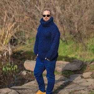 May include: A man wearing a blue turtleneck sweater, blue jeans, and yellow sneakers. He is standing in a wooded area with a rocky path.