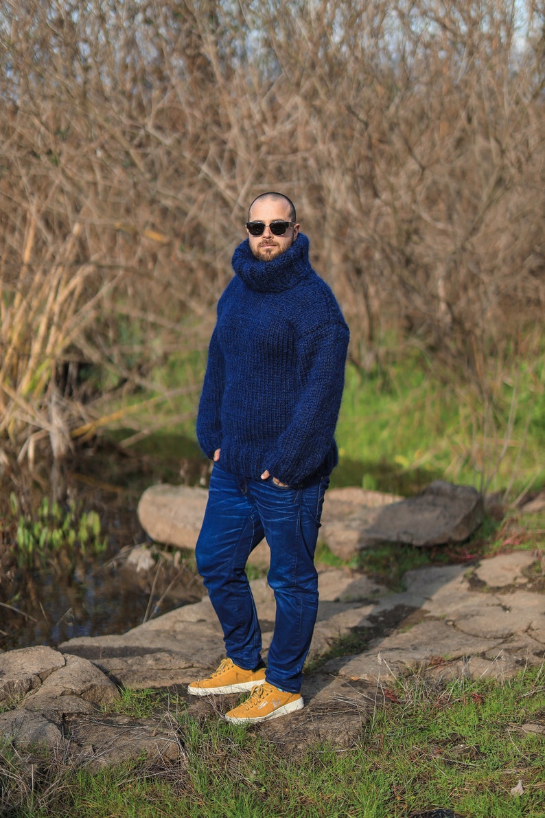 May include: A man wearing a blue turtleneck sweater, blue jeans, and yellow sneakers. He is standing in a wooded area with a rocky path.