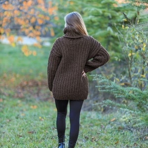 May include: A brown, oversized turtleneck sweater with a chunky knit design. The sweater is paired with black leggings and black sneakers. The person is standing in a grassy area with trees in the background.