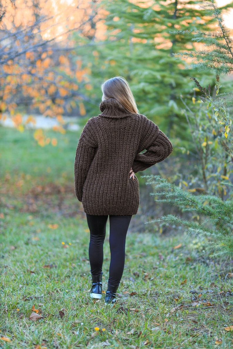 May include: A brown, oversized turtleneck sweater with a chunky knit design. The sweater is paired with black leggings and black sneakers. The person is standing in a grassy area with trees in the background.