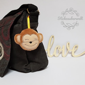 Może przedstawiać: Brązowa torba z płótna z czarno-czerwonym wzorem paisley. Do torby przymocowany jest brązowy pluszowy brelok w kształcie głowy małpy, przymocowany żółtą wstążką. W tle widoczny jest tekst "Stickzauberwelt".