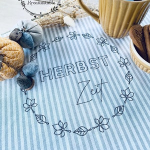 May include: A still-life image features a tan mug with tea, a striped blue and white placemat with the words "HERBST Zeit" embroidered in black, and decorative pumpkins. A small bowl of biscuits and a spoon are also visible.