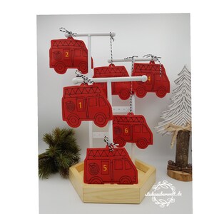 May include: Red felt fire truck ornaments with black stitching and white and black twine hangers. Each ornament is numbered 1 through 6. The ornaments are displayed on a white rack with a wooden base, and a decorative white tree is in the background.