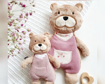 May include: Two plush brown bear toys wearing pink overalls with floral accents. The larger bear is standing and the smaller bear is sitting. A wooden heart-shaped teething ring is in the foreground.