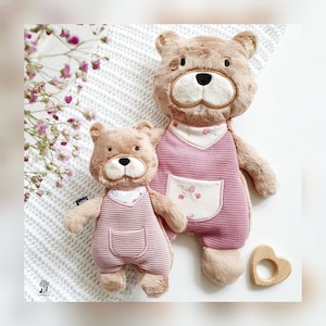 May include: Two plush brown bear toys wearing pink overalls with floral accents. The larger bear is standing and the smaller bear is sitting. A wooden heart-shaped teething ring is in the foreground.