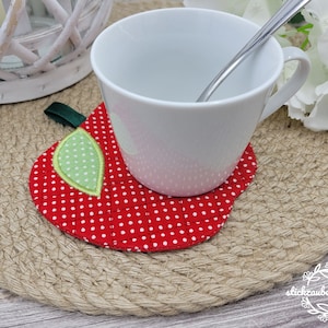 Può includere: Un sottobicchiere di stoffa rosso a pois bianchi a forma di mela con una foglia verde. Una tazza di ceramica bianca con un cucchiaio all'interno è appoggiata sul sottobicchiere.