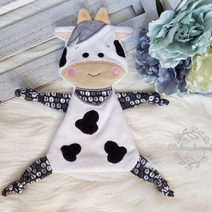 May include: A white and black cow-themed plush toy with a grey nose and pink cheeks. The toy has black and white patterned arms and legs. The toy is lying on a white fluffy surface.