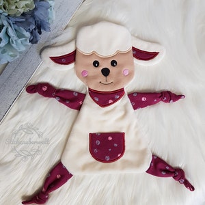Puede incluir: Un cordero de peluche blanco con mejillas rosadas y un patrón de lunares burdeos y blancos. El cordero tiene un bolsillo en el vientre y brazos y piernas anudados.