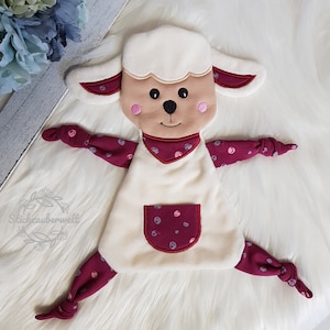 May include: A white plush lamb with a burgundy and white patterned body. The lamb has a red pocket on its belly and a stitched-on face with pink cheeks.