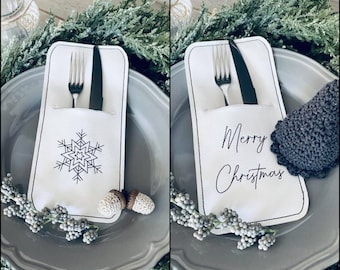 Puede incluir: Dos porta tarjetas de lugar blancos con costuras negras. Uno tiene un diseño de copo de nieve y el otro dice "Feliz Navidad".