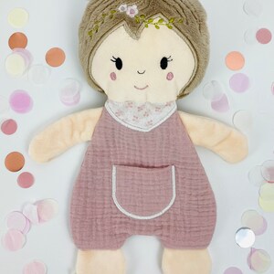 K&ouml;nnte beinhalten: Eine weiche, rosafarbene Pl&uuml;schpuppe mit braunen Haaren, einem wei&szlig;en gestickten Gesicht und einer wei&szlig;en gestickten Tasche auf der Vorderseite ihres rosafarbenen Stramplers.