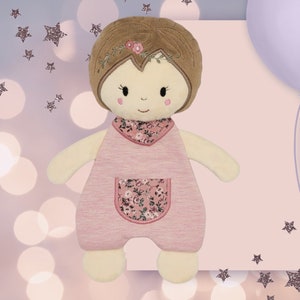 Könnte beinhalten: Eine rosafarbene und cremefarbene Plüschpuppe mit braunen Haaren und einem Kleid mit Blumenmuster. Die Puppe hat eine Tasche auf der Vorderseite ihres Kleides.