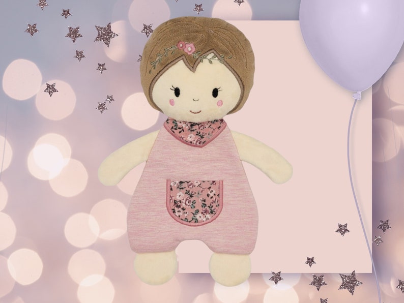 K&ouml;nnte beinhalten: Eine rosafarbene und cremefarbene Pl&uuml;schpuppe mit braunen Haaren und einem Kleid mit Blumenmuster. Die Puppe hat eine Tasche auf der Vorderseite ihres Kleides.