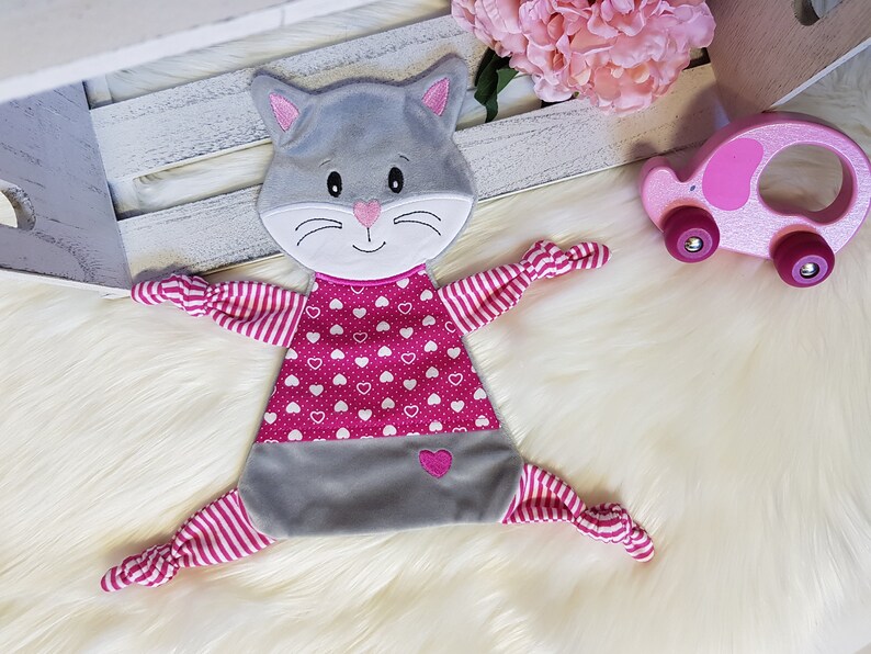 May include: A gray and pink cat-shaped security blanket with a pink heart pattern and pink and white striped arms.