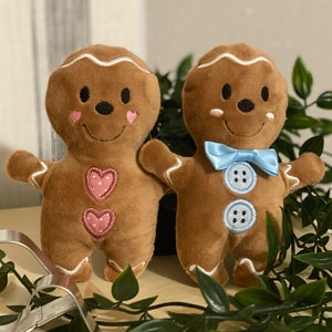 May include: Two plush gingerbread people toys. One is brown with pink hearts and the other is brown with a blue bow and blue buttons.