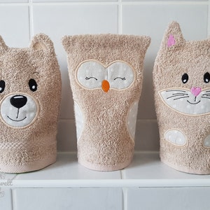 May include: Three beige washcloths shaped like animals: a bear, an owl, and a cat. The bear has a black nose and eyes, the owl has an orange beak, and the cat has pink ears. Each washcloth has embroidered details and is made of a soft, absorbent material.