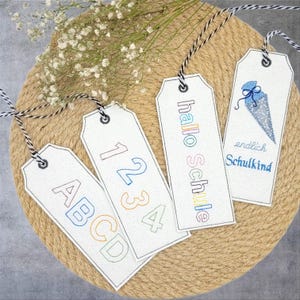 Peut inclure: Quatre étiquettes cadeaux blanches avec des broderies colorées. Les étiquettes présentent les mots "hallo Schule", "endlich Schulkind", "1234" et "ABCD".