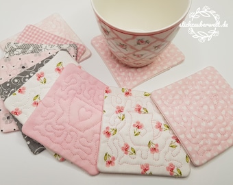 May include: Set of pink and gray fabric coasters with various floral and geometric patterns. The coasters are quilted and feature a delicate stitching detail. The coasters are arranged in a stack with a cup and saucer on top.