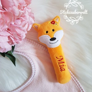 以下が含まれることがあります： 白い顔と頭に赤い花が付いたオレンジ色のぬいぐるみキツネのガラガラ。ガラガラには「ミア」という名前が刺繍されています。