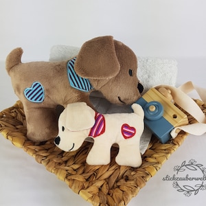 Peut inclure: Deux chiens en peluche, l'un brun et l'autre crème, avec des cœurs bleus et rouges sur la poitrine, sont assis dans un panier en osier avec un appareil photo jouet en bois et une sangle blanche.