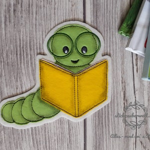 Op de afbeelding: Een groene viltworm met een bril leest een geel boek. De worm glimlacht en heeft een gelukkige uitdrukking. Het boek is open en de pagina's zijn zichtbaar. De worm zit op een houten oppervlak.