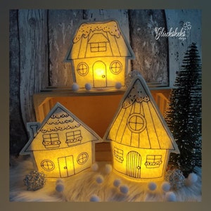 Könnte beinhalten: Drei weiße Filzhäuser mit schwarzen Umrissen und Fenstern, von innen mit warmweißen Lichtern beleuchtet. Die Häuser stehen auf einer Holzkiste mit weißen Pompons, die um sie herum verstreut sind. Der Text "Glückskeks design" ist in der oberen rechten Ecke des Bildes sichtbar.