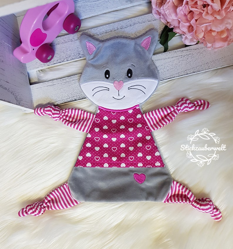 May include: A gray and pink cat-shaped baby toy with a pink heart pattern on the front and pink and white striped arms and legs. The toy has a knotted end on each arm and leg.