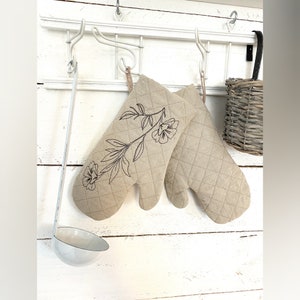 以下が含まれることがあります： ベージュのオーブンミット2枚組。片方のミットには花柄の刺繍が施されています。ミットは白い金属製のラックに掛けられています。