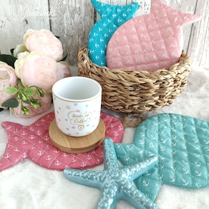 May include: A set of four teal and pink fish-shaped pot holders with white anchor designs. A white mug with colorful polka dots and the text "Wake up and smell the coffee" sits on a wooden coaster on top of a pink fish pot holder. A teal starfish sits on top of the pot holders.