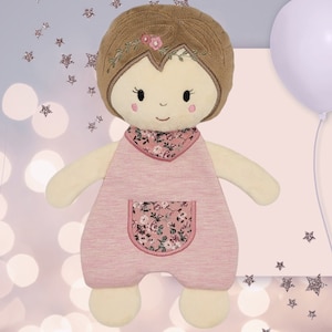 K&ouml;nnte beinhalten: Eine rosafarbene Pl&uuml;schpuppe mit braunen Haaren, einem Kleid mit Blumenmuster und einer wei&szlig;en gestickten Tasche. Die Puppe hat ein freundliches Gesicht mit schwarzen Augen und einem rosafarbenen L&auml;cheln.