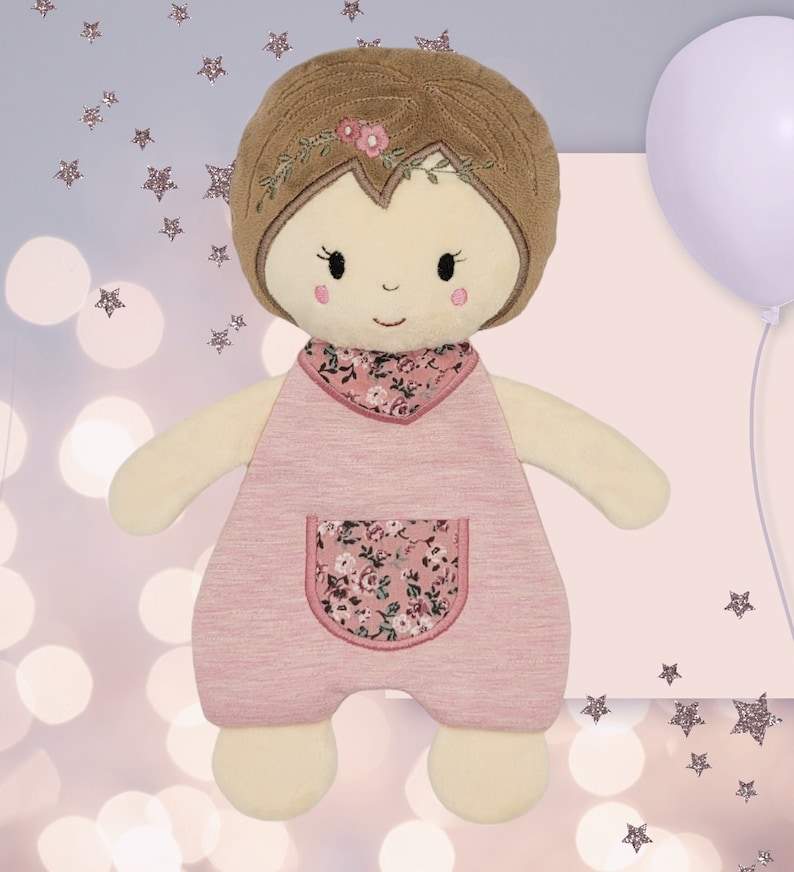 K&ouml;nnte beinhalten: Eine rosafarbene Pl&uuml;schpuppe mit braunen Haaren, einem Kleid mit Blumenmuster und einer wei&szlig;en gestickten Tasche. Die Puppe hat ein freundliches Gesicht mit schwarzen Augen und einem rosafarbenen L&auml;cheln.