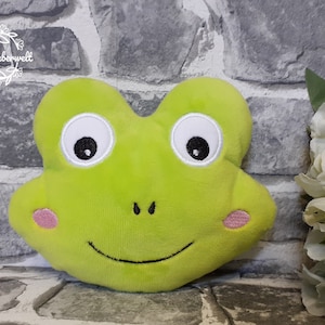 Puede incluir: Cabeza de rana de peluche verde lima con ojos blancos grandes, pupilas negras y mejillas rosas. La rana tiene una sonrisa y fosas nasales negras cosidas. El texto "Stichzauberwelt" está en la esquina superior izquierda.