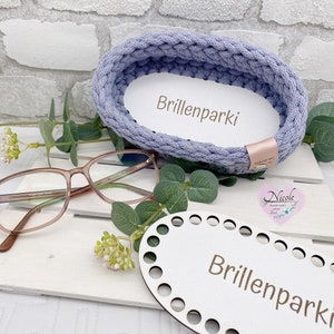 Può includere: Un cestino in filo intrecciato blu chiaro con la scritta "Brillenparki" su un inserto bianco. Sono presenti anche occhiali con montatura marrone e una base in legno con la stessa scritta. Il cestino è su una superficie bianca.