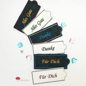 Könnte beinhalten: Sechs Filz-Etiketten mit verschiedenen Grüßen auf Deutsch. Die Etiketten sind weiß mit schwarzer, blauer, grüner, goldener und roter Stickerei. Die Grüße lauten "Alles Gute", "Danke" und "Für Dich".