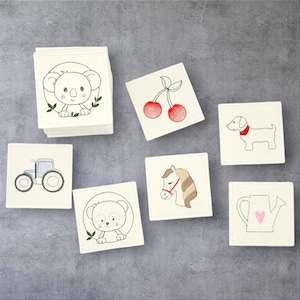 Könnte beinhalten: Ein Set mit 10 weißen Stoffquadraten mit verschiedenen Stickereien. Die Designs beinhalten einen Koala, Kirschen, einen Hund, ein Pferd, einen Bären, einen Traktor und eine Gießkanne.