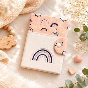 May include: A cream-colored notebook with a rainbow design embroidered on the front. The top flap is peach-colored with a quilted pattern of rainbows and clouds. A small peach-colored flap with a button closure is on the right side. A wooden cloud-shaped toy and a pink heart are also in the image.