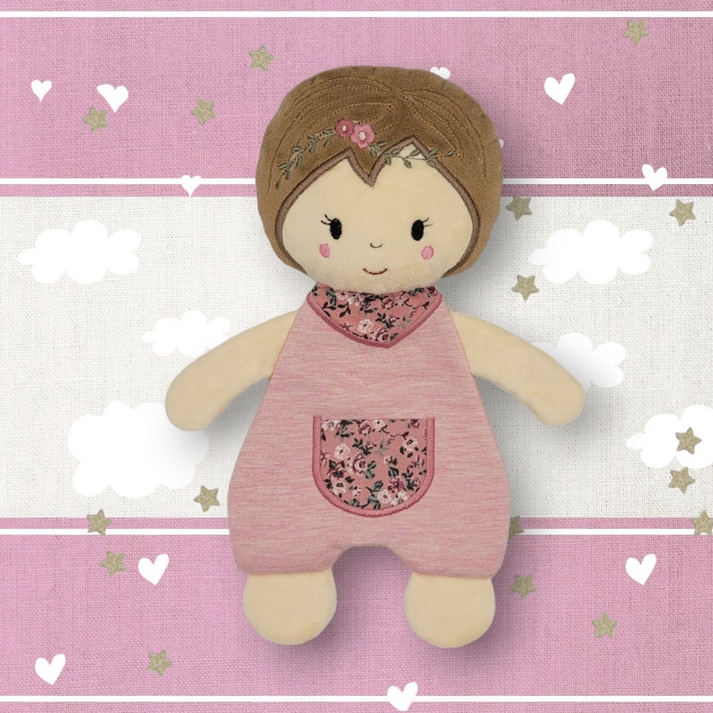 K&ouml;nnte beinhalten: Eine rosafarbene und braune Pl&uuml;schpuppe mit einem Kleid mit Blumenmuster und einem Stirnband. Die Puppe hat ein l&auml;chelndes Gesicht und braunes Haar.