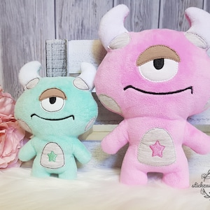 May include: Two plush one-eyed monster toys. The larger monster is pink with white horns and a brown eye. The smaller monster is teal with white horns and a brown eye. Both monsters have a white star on their bellies.