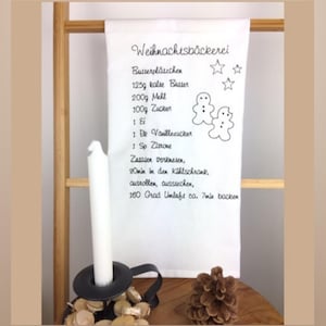 Könnte beinhalten: Ein weißes Küchentuch mit einem schwarzen gestickten Rezept für Lebkuchen. Das Rezept enthält Zutaten wie Butter, Mehl, Zucker und Vanilleextrakt. Das Tuch zeigt auch zwei Lebkuchenmann-Illustrationen.