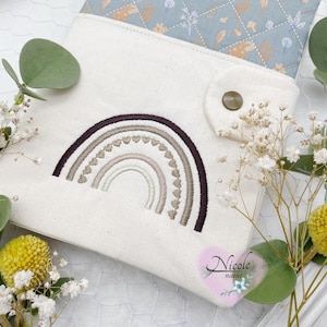 Può includere: Una pochette in tela bianca con un arcobaleno marrone, beige e bianco ricamato sul davanti. La pochette ha una patta con una chiusura a bottone marrone. Il testo "Nicole macht" è ricamato nell'angolo in basso a destra della pochette.