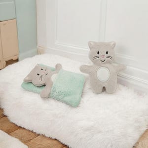 Puede incluir: Un juguete de peluche gris con detalles bordados y una barriga blanca. Junto a él, un juguete de gato a juego en un juego de almohada y manta verde menta. Los juguetes están sobre una alfombra blanca y esponjosa.