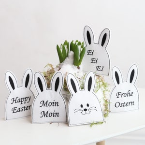 May include: Five white felt bunny-shaped Easter decorations with black embroidered details. The bunnies have different greetings, including "Happy Easter", "Moin Moin", "Ei Ei Ei", and "Frohe Ostern".