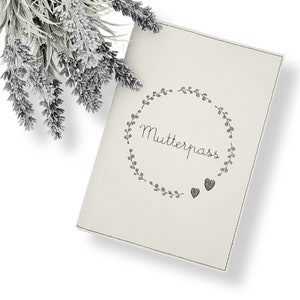 Könnte beinhalten: Cremefarbenes Journal mit dem Wort "Mutterpass" in Schriftform in einem floralen Kranz und zwei Herzformen. Das Journal hat graue Nähte an den Rändern. Ein Zweig Lavendel befindet sich in der oberen linken Ecke.