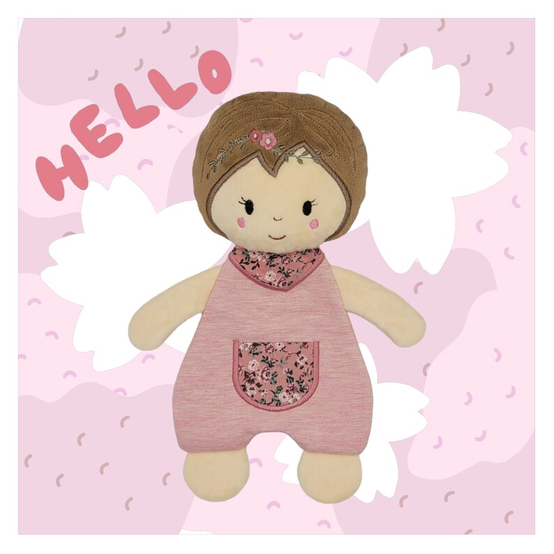 K&ouml;nnte beinhalten: Eine rosafarbene und braune Pl&uuml;schpuppe mit gestickten Blumendetails. Die Puppe hat braunes Haar, ein rosafarbenes Kleid und eine Tasche mit Blumenmuster. Die Puppe sitzt auf einem rosa und wei&szlig;en Hintergrund mit dem Wort "HELLO" in rosa.